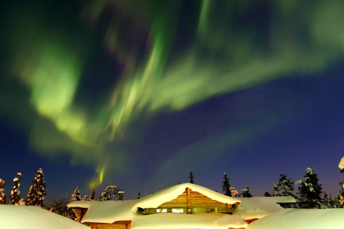 Yellow-green northern lights arching over snow-covered cabins with a lit window