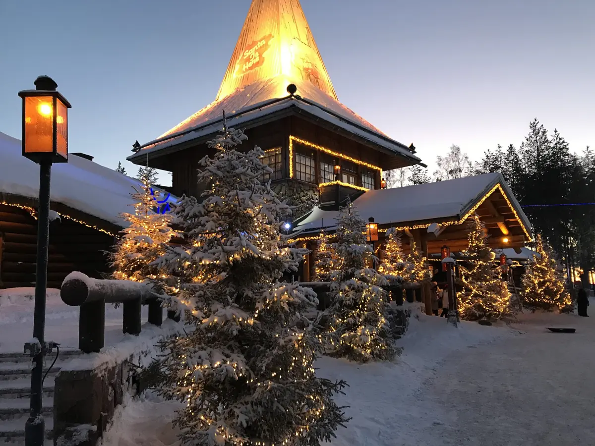 Snow-covered Santa Claus Village at dusk with glowing roof and Christmas lights